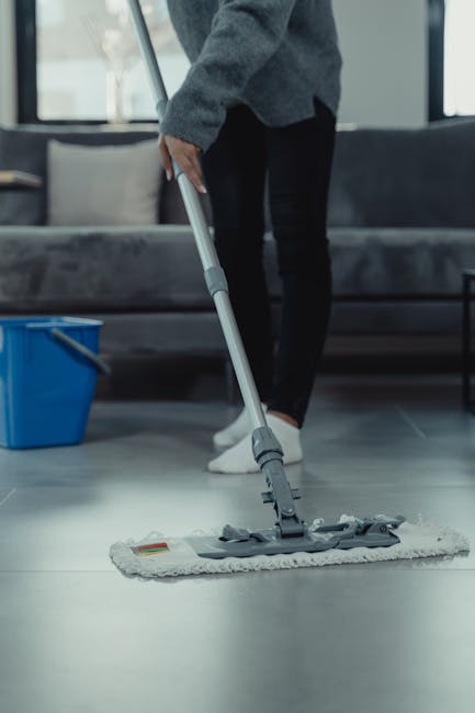 A team of three professional cleaners standing in a modern living room with light-colored tiled flooring, white walls, and large windows. The cleaners are dressed in dark uniforms with aprons, holding