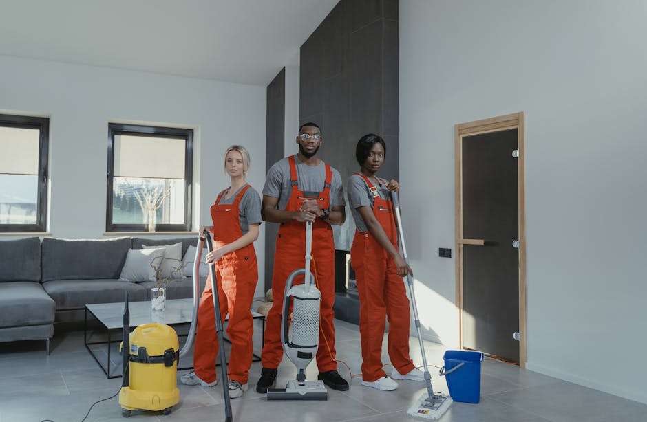 Three professional cleaners from Ealing Cleaner dressed in grey shirts and orange overalls are performing surface cleaning and deep cleaning tasks in a modern living room. The room features a grey sec