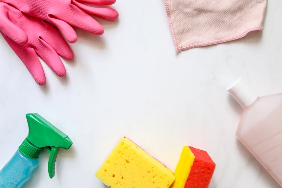 An arrangement of cleaning tools and supplies on a white surface, including a pair of pink rubber gloves, a green spray bottle, a yellow and red cleaning sponge, a pink cloth, and a partially visible
